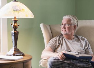 A senior man at home, sitting in the living room reading a book by the light of a table lamp.